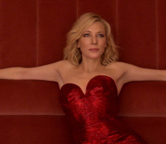 A woman in a strapless red sequined dress sits elegantly on a dark red couch, her arms resting on the backrest, exuding confidence and grace.