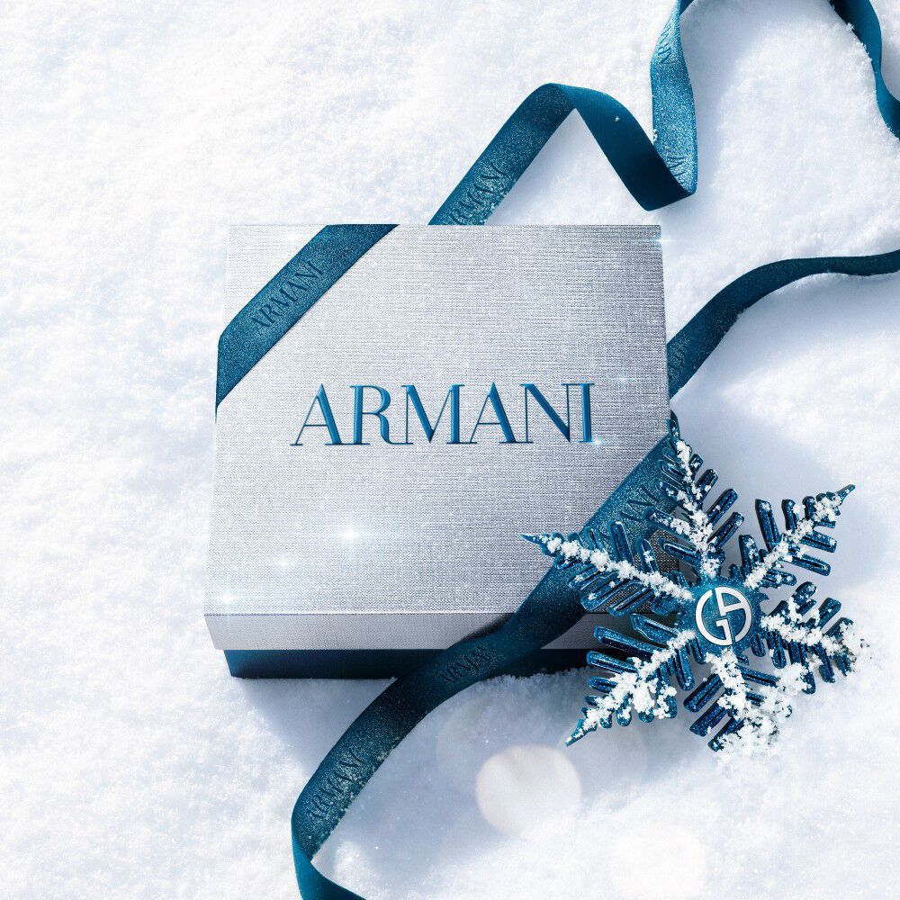 Armani beauty gift box — silver gift box wrapped with blue satin ribbon and a metallic snowflake ornament, resting on fresh white snow.