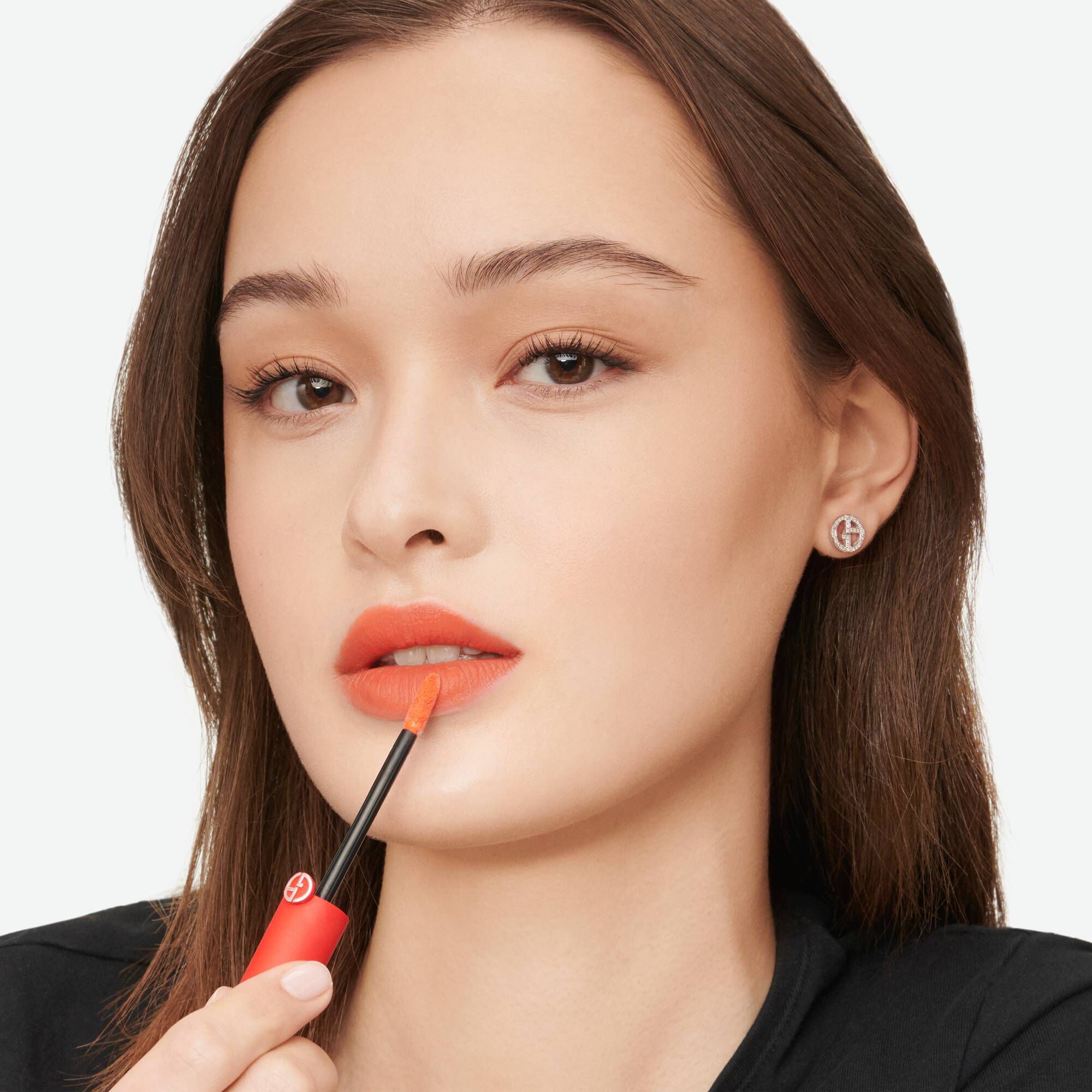 A young woman applying coral lipstick using a small brush from a red tube, with subtle makeup and simple jewelry.