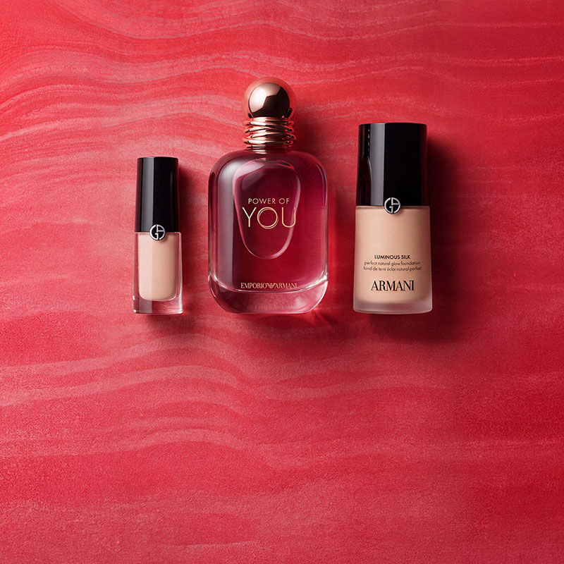 Une pochette rouge Armani Beauty entouree de produits de parfum, de maquillage et de soin sur un fond rouge texture.