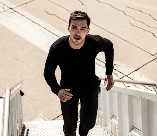 A man dressed in black runs up a set of metal stairs, casting a shadow on the ground. The background features a bright outdoor setting.