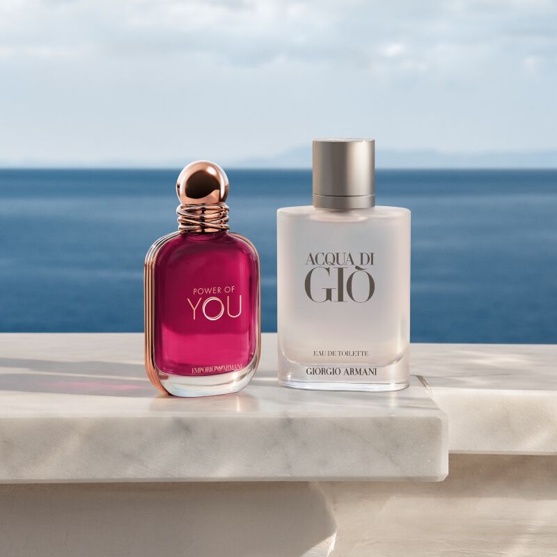 Deux flacons de parfum Giorgio Armani, « Power of You » d'Emporio Armani (bouteille rose fonce) et « Acqua di Gio » (bouteille depolie), poses sur un rebord en marbre blanc avec la mer bleue et le ciel en arriere-plan.