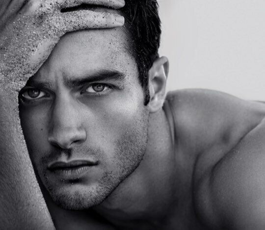 A close-up black and white portrait of a young man with a serious expression, resting his forehead on his arm while his skin is dusted with fine sand.
