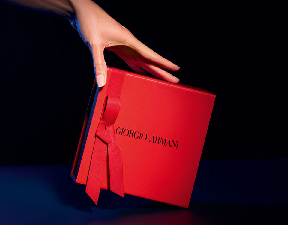 A hand reaches for a red gift box tied with a ribbon, featuring the name "Giorgio Armani" on the front. The background is dark, highlighting the box.