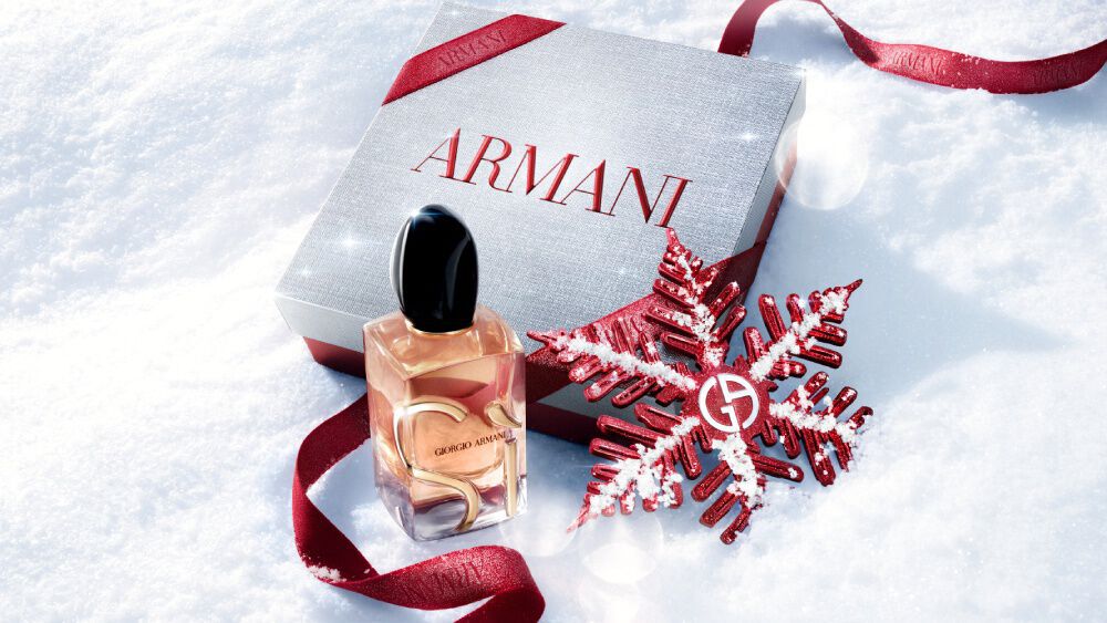 Si Eau de Parfum by Giorgio Armani, gold-toned bottle resting in snow with a red glitter ribbon and Giorgio Armani snowflake ornament — women's fragrance holiday gifting visual.