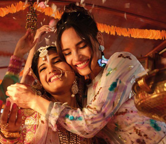 Two joyful women embrace each other, smiling brightly amidst a festive setting, adorned in elaborate traditional attire and jewelry, with confetti or flower petals falling around them.
