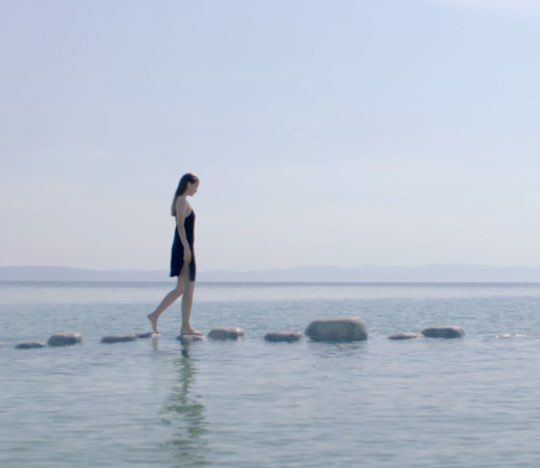 A woman in a black dress walks barefoot across stones in shallow water, with a serene lake and distant mountains in the background under a clear blue sky.
