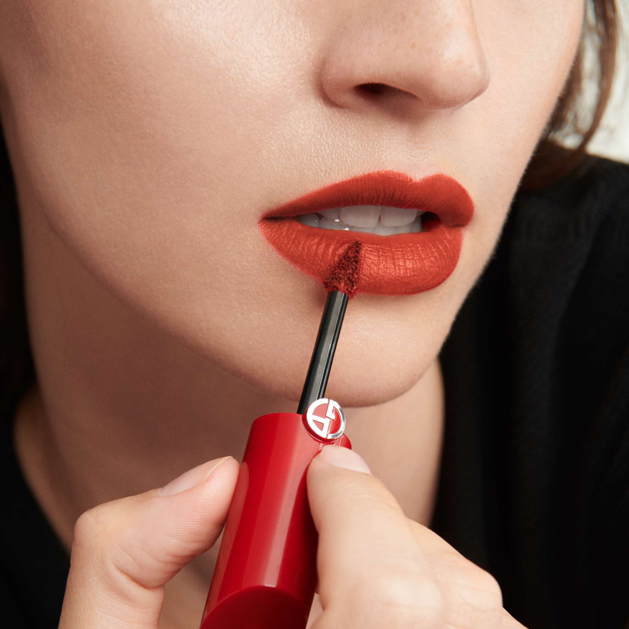 A close-up of a person's mouth as they apply a bold red lip color using a wand applicator. The person has a neutral expression and is wearing a dark top.