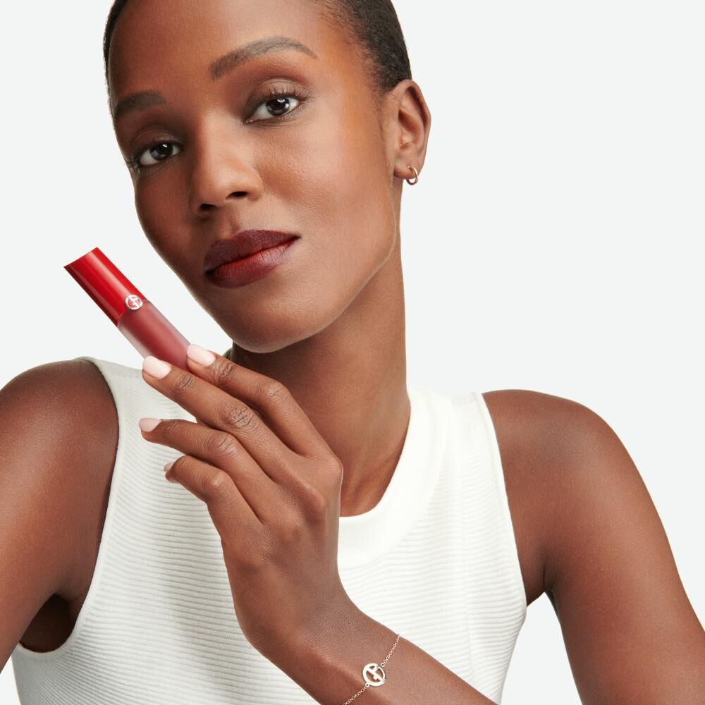 A woman with short hair wears a white sleeveless top and holds a red lipstick in one hand, showcasing a deep maroon lip color. She looks confidently at the camera, wearing minimal jewelry.