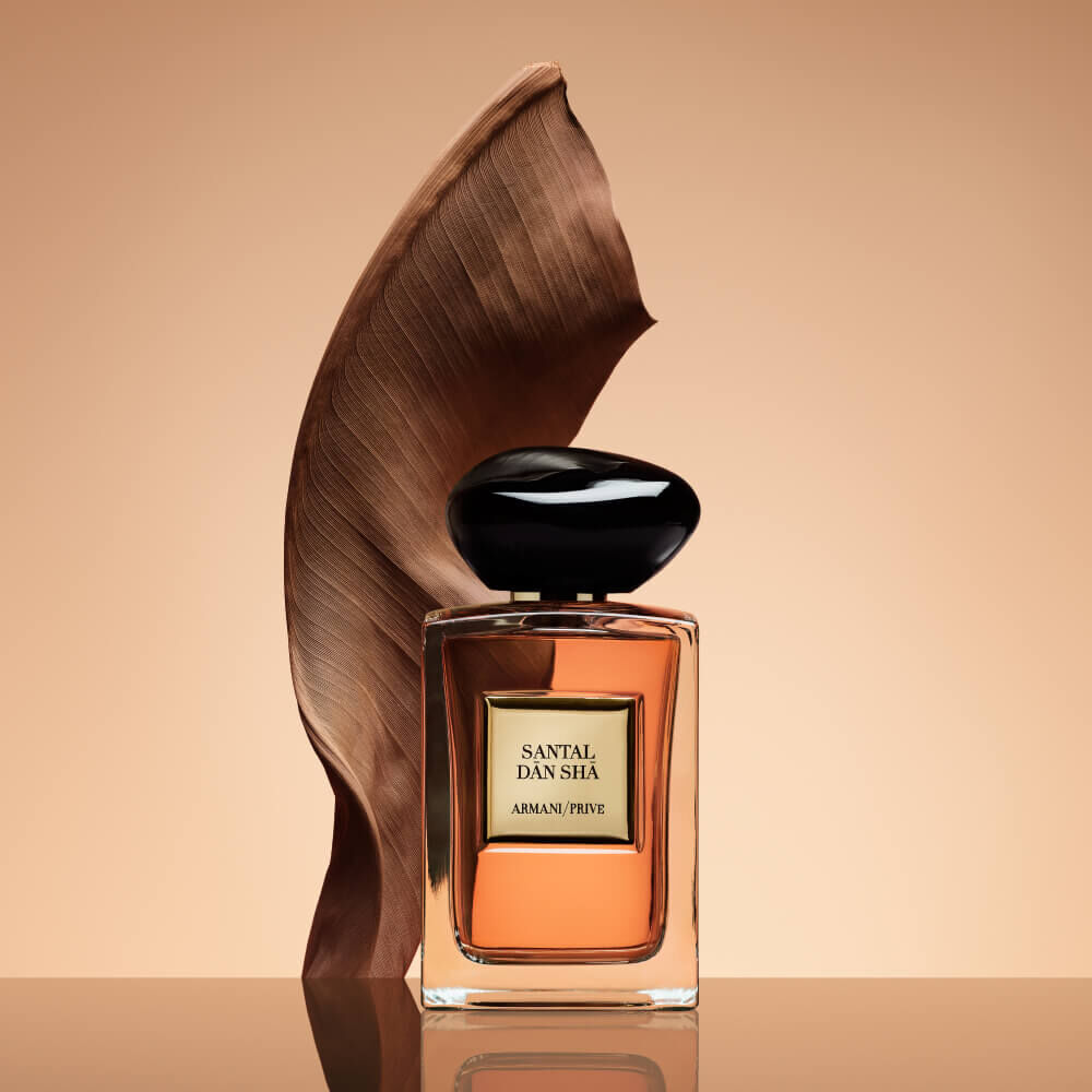 Armani Prive Les Eaux Santal Dān Shā eau de parfum, transparent rectangular glass bottle with black rounded cap, set against a warm terracotta and bronze sculptural background.