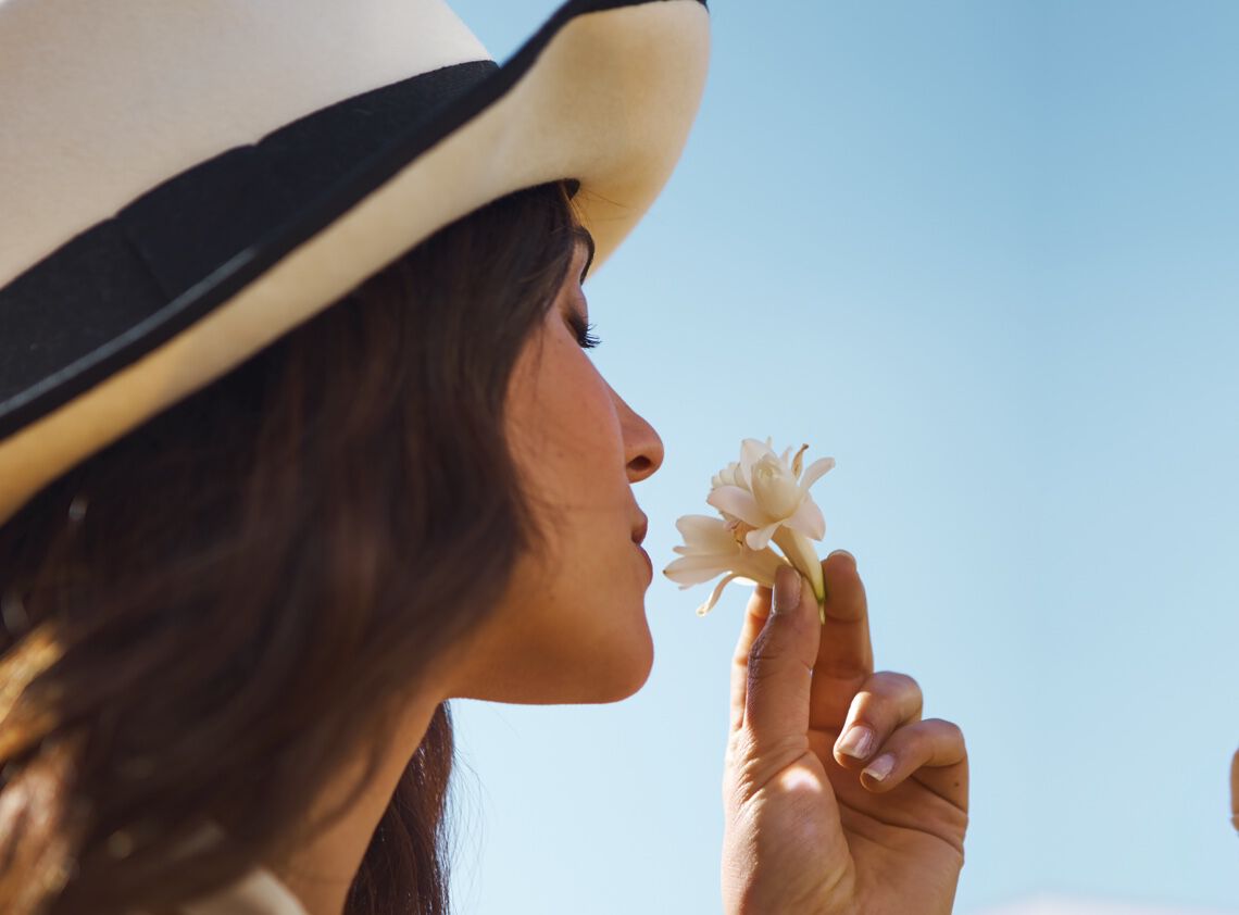 A woman wearing a wide-brimmed hat gently holds a small white flower to her nose, enjoying its scent against a clear blue sky.
