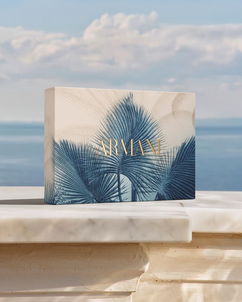 Un coffret cadeau Armani orne d'un motif botanique bleu, pose sur du marbre face a la mer. Un hommage a la collection "Armani Mare", melant savoir-faire couture et elegance solaire pour un rituel de beaute d'exception.