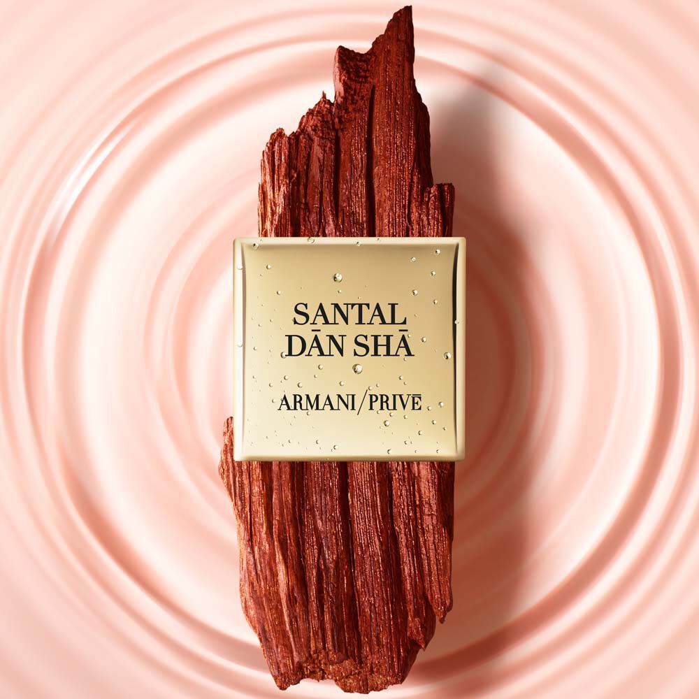 A bottle of Armani Prive Santal Dan Sha perfume displayed on a textured wooden background, with water droplets on the bottle reflecting a soft pink surface.