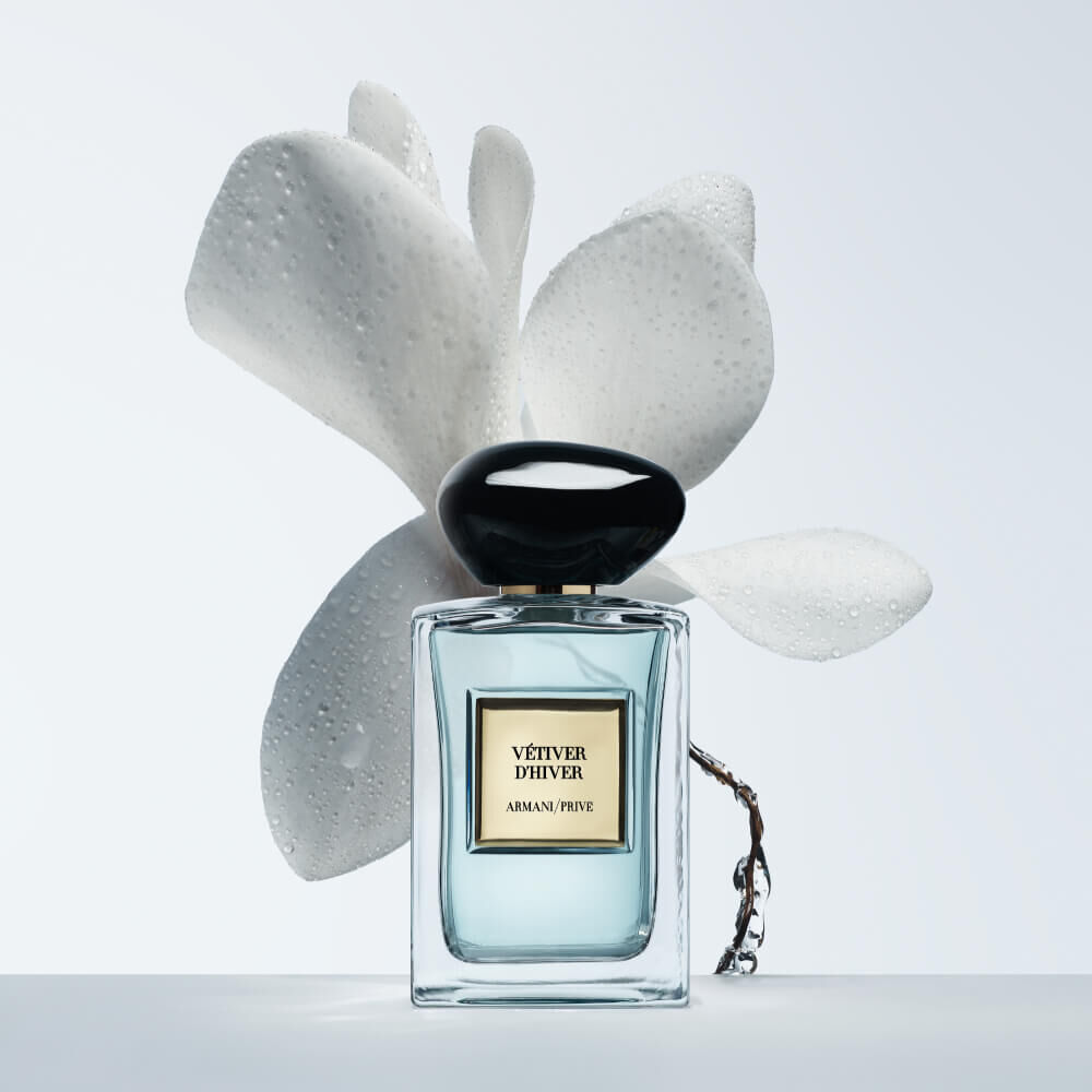 Armani Prive Les Eaux Vetiver d'Hiver eau de parfum, transparent rectangular glass bottle with black rounded cap, beside a sculptural white flower on a pale grey-blue background.