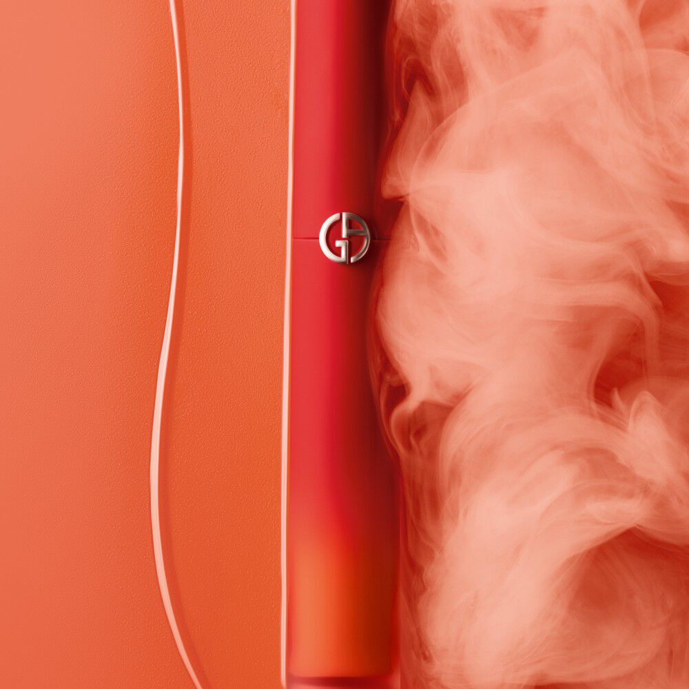 A vibrant red cosmetic tube partially obscured by soft, swirling smoke against an orange background. The tube features a silver logo.