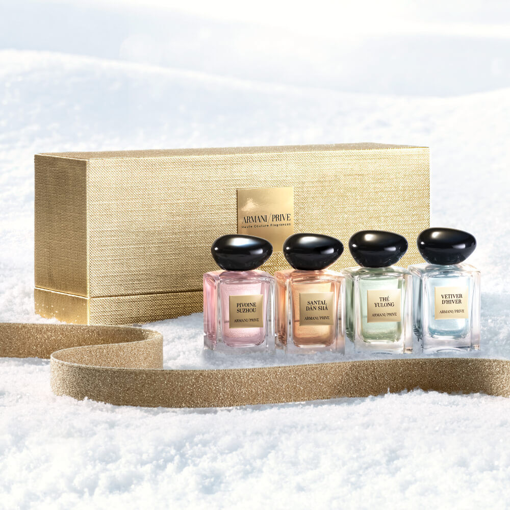 Armani Prive Les Eaux Holiday miniature gift set: gold rectangular box alongside four miniature eau de parfum bottles, photographed on a pale textured snow surface.