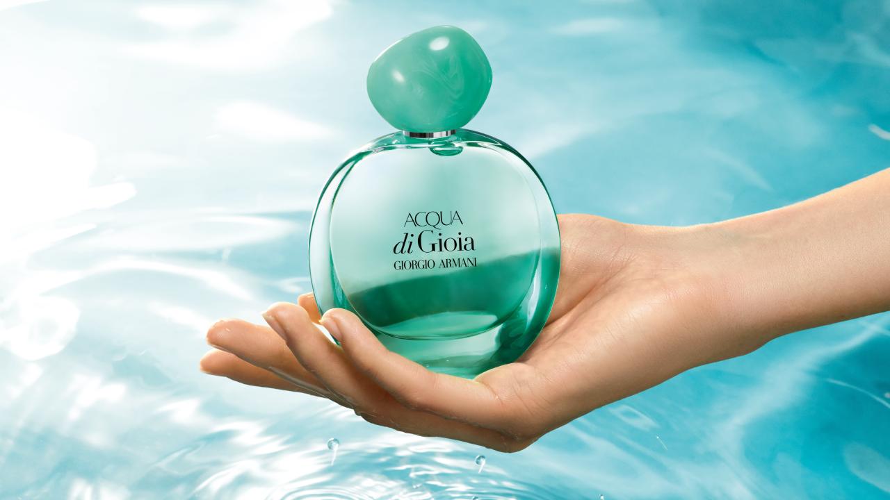 A hand holding a bottle of Acqua di Gioia perfume by Giorgio Armani against a sparkling blue water background.