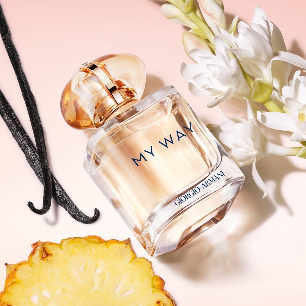 Armani My Way perfume bottle displayed with vanilla beans, white tuberose flowers, and a slice of pineapple on a soft pink background, emphasizing its natural fragrance ingredients.